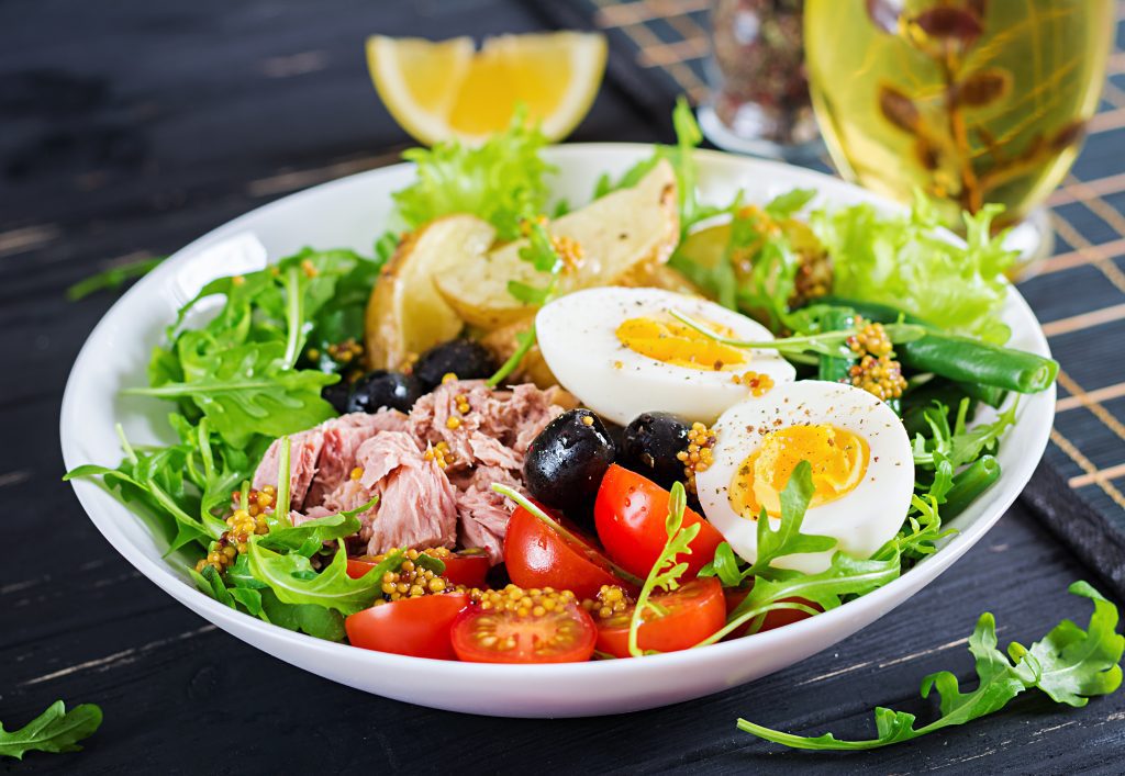 Salade copieuse avec saine de thon, haricots verts, tomates, œufs, pommes de terre, gros plan d'olives noires dans un bol sur la table.